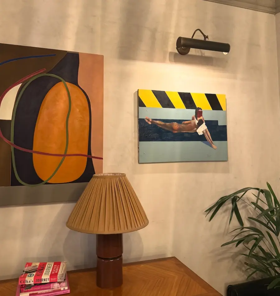 Two paintings above a pleated table lamp, books and a palm on a wooden sideboard
