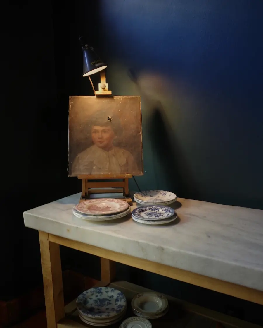 An old child's portrait on a small easel lit by a clamp lamp, with china plates on a marble table